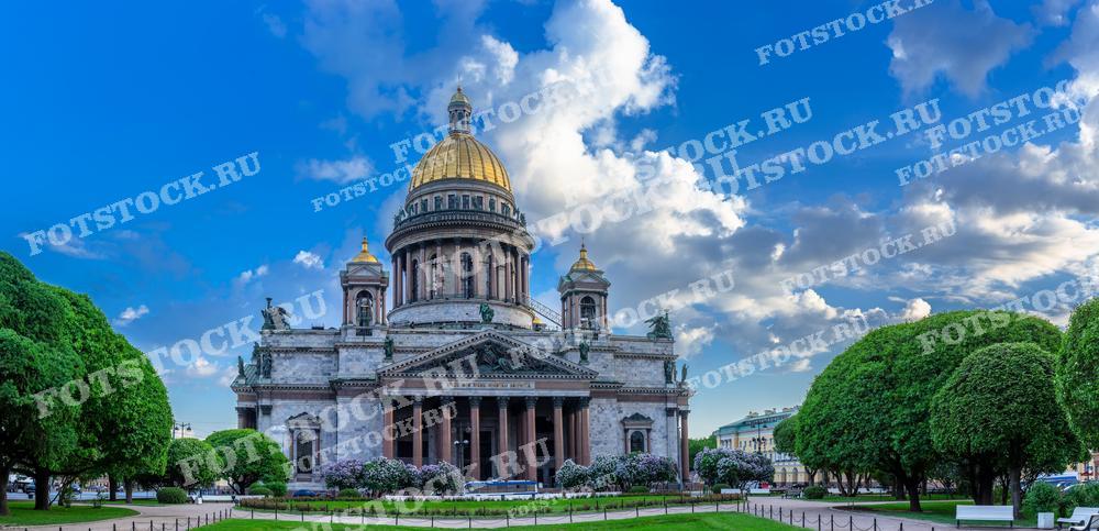 На фотографии изображен Исаакиевский собор в Санкт-Петербурге, окруженный зелеными деревьями и голубым небом. Фото может быть использовано для туристических и образовательных целей, а также для иллюстраций в СМИ и рекламных материалов.